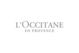 A imagem mostra o logotipo da L'Occitane en Provence em um fundo branco. O texto está escrito em uma fonte serifada cinza simples, com "L'OCCITANE" em letras maiores acima de "EN PROVENCE".